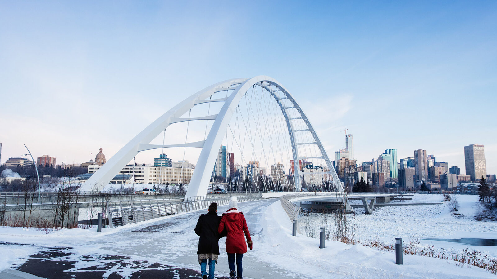 Je ziet twee vrouwen die bij de grote brug van Edmonton lopen. Overal ligt sneeuw en de lucht is helderblauw.